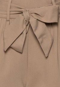 Cintura in tessuto beige annodata a un fiocco, fissata a pantaloni su misura. Texture liscia con un leggero lucido. La vita è piatta, con pieghe sottostanti.