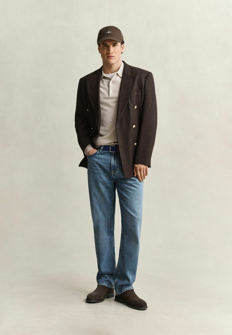 Young man wearing a dark brown blazer, beige shirt, blue jeans, brown shoes, and a brown baseball cap standing with one hand in pocket.