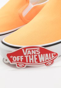 Chaussures slip-on en toile orange vif avec une semelle en caoutchouc blanche, ornées d'une bande noire et d'une étiquette rouge "Vans Off the Wall".