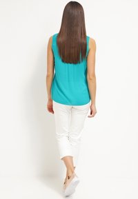 Turkusowy top bez rękawów o gładkiej teksturze, zestawiony z białymi spodniami 3/4. Modelka odwrócona, ukazuje długie włosy i casualowe obuwie.