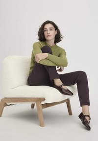 Vrouw met kort krullend haar zit met gekruiste benen op een witte textuurstoel, gekleed in een olijfgroene trui, donkere broek en donkere gespschoenen.