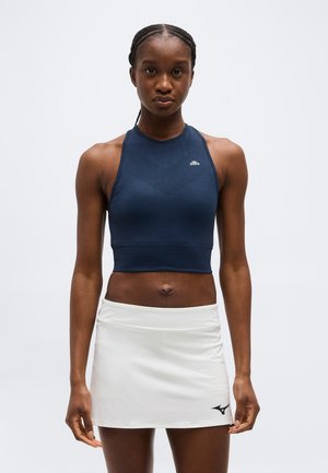 Sportliche Frau steht mit Blick nach vorne, trägt ein ärmelloses marineblaues Crop-Top und weißen Sportrock vor einem schlichten Hintergrund.