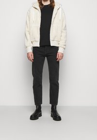 Creme faux fur zip-up hoodie med hvide snore, parret med en sort skjorte, sorte jeans og sorte chelsea boots. Ensfarvet baggrund.