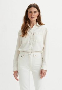 Camisa de denim blanca con cuello puntiagudo, cierre de botones y dos bolsillos en el pecho. Llevada con pantalones de denim blancos a juego. Textura lisa.