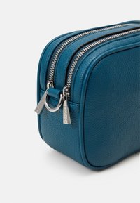 Kosmetisk väska i blågrönt läder med strukturerad yta, utrustad med två silverdragkedjor och metalliska detaljer för enkel åtkomst.