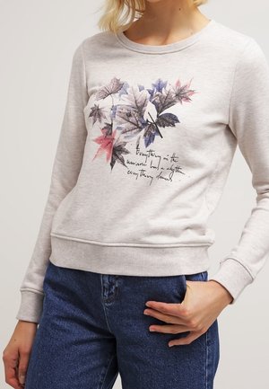 Sweat-shirt gris clair avec un motif coloré de feuille et un texte en cursive porté par une personne avec une main posée sur un jean bleu.