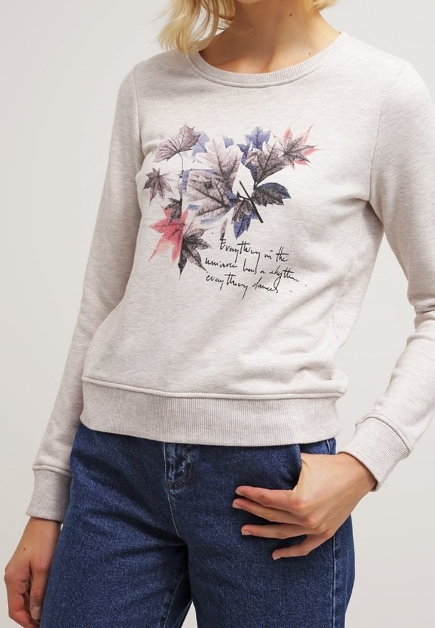 Sweat-shirt gris clair avec un motif coloré de feuille et un texte en cursive porté par une personne avec une main posée sur un jean bleu.