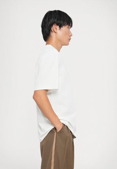 T-shirt bianco a maniche corte con colletto rotondo, texture liscia e vestibilità rilassata, abbinato a pantaloni marroni con strisce laterali.
