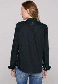 Maglietta nera a maniche lunghe con colletto, piega sul retro e dettaglio ricamato a forma di ancora. Il tessuto è liscio con un motivo contrastante all'interno dei polsini.