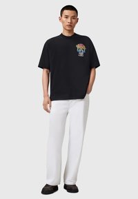 T-shirt en coton noir à manches courtes, avec un graphique de crâne coloré sur la poitrine. Associé à un pantalon blanc à jambes droites et des chaussures sombres.