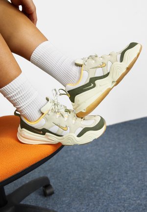 Masivne sneakersy z beżową siateczką na górze, oliwkowozielonymi akcentami, żółtymi detalami i fakturowaną kremową podeszwą. Widoczne białe prążkowane skarpetki.