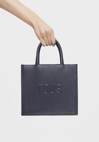 Bolso de piel navy con superficie texturizada y logotipo "TOUS" grabado. Presenta asas cortas y una forma rectangular estructurada.