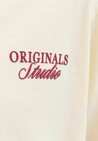 Material de algodón color crema con texto bordado en color marrón que dice "ORIGINALS Studio". Textura suave, diseño minimalista.