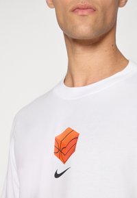 Biela bavlnená tričko s grafikou oranžovej basketbalovej kocky a čiernym logom Nike pod ňou. Okruhly výstrih a krátke rukávy.