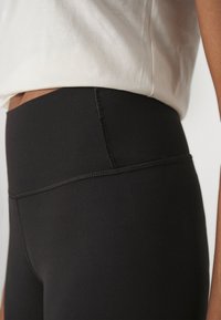 Leggings neri a vita alta realizzati in materiale elasticizzato con una texture liscia, dotati di una vita piatta e sottili dettagli di cucitura.