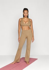 Μπραζίνα σπορ σε ταμπά απόχρωση και ταιριαστά κολάν με φλάρισμα, κατασκευασμένα από απαλό ύφασμα. Το μοντέλο στέκεται σε ένα ροζ στρώμα γιόγκα με γυμνά πόδια. Απλός σχεδιασμός, χωρίς μοτίβα.