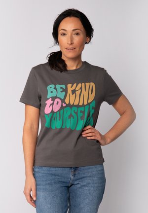 Frau mit dunklem Haar im Pferdeschwanz, trägt ein graues T-Shirt mit buntem Schriftzug "Sei freundlich zu dir selbst" und blaue Jeans, steht mit einer Hand in der Hüfte.