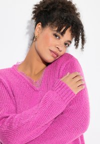Pull en V de couleur rose vif. Texture en tricot épais avec des poignets côtelés. Bordure en dentelle au niveau du col. Apparence douce et chaleureuse.