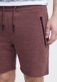 Shorts bourgogne en tissu doux, avec une taille à cordon de serrage et une poche zippée noire sur le côté.