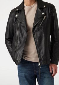 Schwarze Leder-Motorradjacke mit silbernem Zubehör, asymmetrischen Reißverschlüssen und einem strukturierten hellgrauen Hemd darunter, getragen mit blauen Jeans.