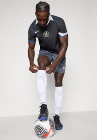Maglietta da calcio nera con dettagli blu, pantaloni corti coordinati con pattern e calze bianche. In piedi su una palla da calcio bianca e nera con dettagli arancioni.