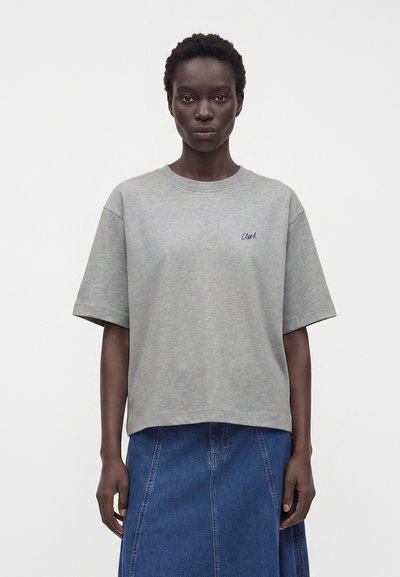 Femme portant un t-shirt gris ample à manches courtes avec un petit texte sur la poitrine et une jupe en jean bleue taille haute sur un fond uni.