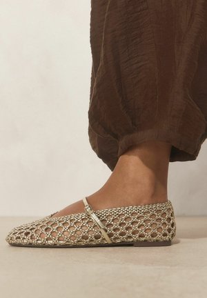 Stopa w złotym plecionym płaskim bucie z paskiem wokół kostki, zestawiona z luźnymi brązowymi spodniami na jednolitym tle.