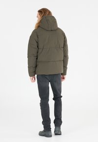 Oliven grøn polstret jakke med hætte, der har en slank silhuet og vandret syninger. Parret med mørkegrå jeans og støvler.
