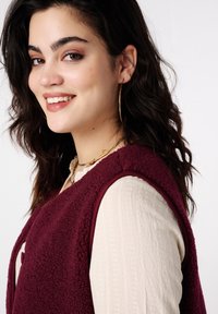 Gilet en polaire bordeaux aux bords arrondis, fermeture par boutons, porté sur un haut crème texturé. Comprend de grandes boucles d'oreilles anneaux et un collier superposé.