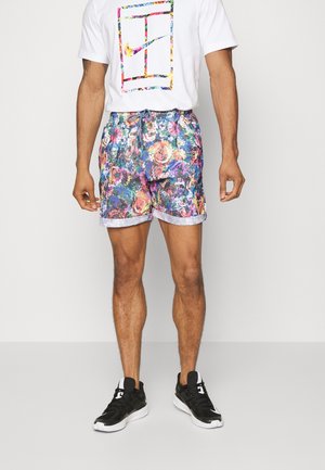 Man die een wit t-shirt draagt met kleurrijk logo, meerkleurige bloemmotief shorts en zwart-witte sportschoenen, staand tegen een effen achtergrond.