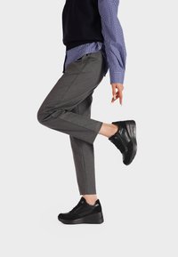 Sneakers neri dal design elegante, realizzati in pelle e suede, con una suola strutturata e sottili dettagli in metallo. Indossati con pantaloni grigi.