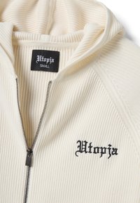 Sudadera con capucha color crema acanalada con cremallera, bordado negro "Utopia" en el pecho y etiqueta a juego en el interior del cuello, talla pequeña.