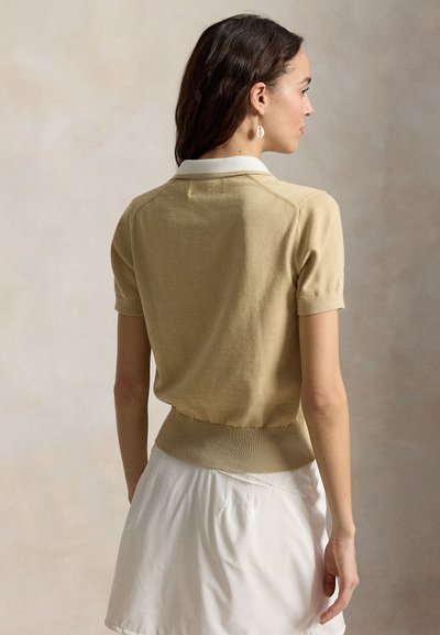 Γυναίκα με σκούρα μαλλιά που φοράει μπεζ πουλόβερ με κοντά μανίκια, λευκό γιακά και λευκή φούστα, στέκεται με την πλάτη σε ουδέτερο φόντο.