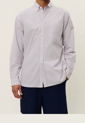 Mann trägt ein langärmeliges weißes Hemd mit dünnen vertikalen roten Streifen und dunkelblaue strukturierte Shorts, eine Hand entspannt, eine in der Tasche.