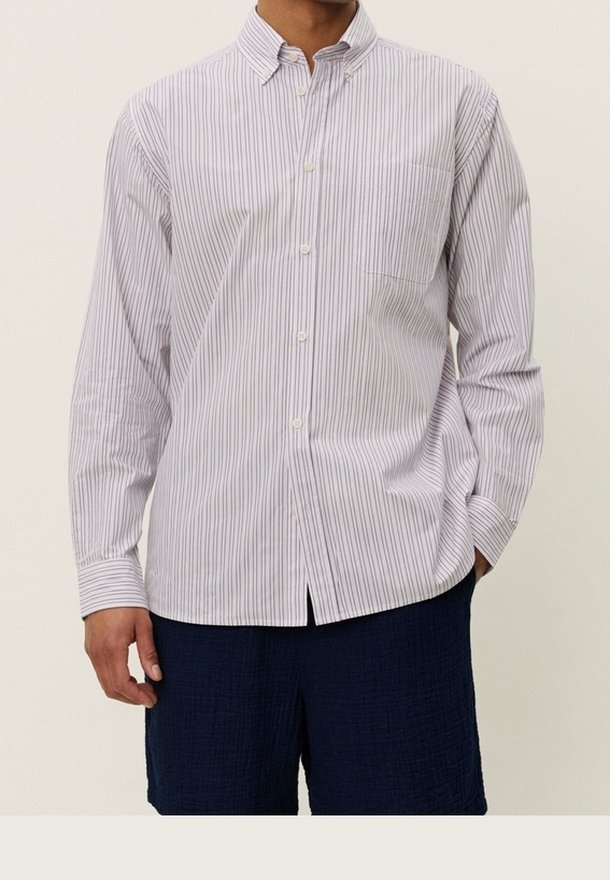 Homme portant une chemise blanche à manches longues avec de fines rayures verticales rouges et un short texturé bleu foncé, une main détendue, l'autre dans la poche.
