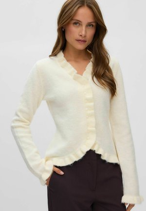 Femme portant un cardigan duveteux couleur crème avec des bordures à volants et des boutons, combiné à un pantalon foncé, sur un fond uni clair.