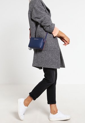 Mörkblå läder crossbody-väska med rektangulär form, guldlogotyp och en kort axelrem. Bärs tillsammans med en grå kappa och svarta byxor.