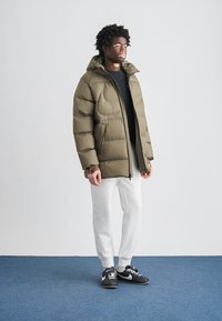 Oljčno zelena puhovka z vbodeno teksturo, z zadrgo po celotni dolžini in kapuco, združena z belimi trenirkami in črnimi supergami.