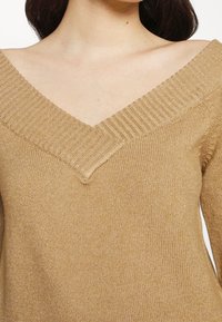 Maglione a maglia beige con scollatura a V e dettagli a coste. La texture appare morbida e il tessuto ha una leggera lucentezza.