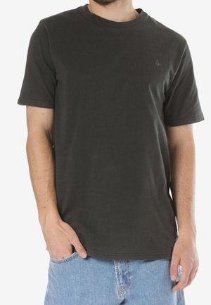 Mann trägt ein schlichtes, dunkelgraues T-Shirt mit Kurzarm und helle blaue Jeans, steht mit einer Hand in der Tasche.