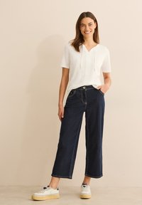 Witte blouse met korte mouwen en een V-hals, gecombineerd met donkerdenim wijde broeken met zichtbare stiksels en zakken. Witte sneakers.