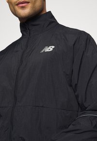 Homme portant une veste noire zippée New Balance avec un col montant visible des épaules au menton.