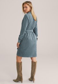 Robe en velours côtelé bleu atténué avec ceinture à nouer, manches longues et texture à côtes verticales, assortie à des bottes en suède beige hautes.