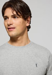 Sudadera de algodón gris con cuello redondo y puños acanalados. Presenta un pequeño logo en azul marino en el pecho. Textura suave.