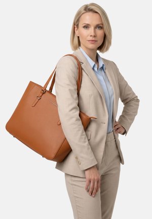 Professionelle Frau im beigen Anzug trägt eine große hellbraune Ledertasche auf der Schulter und steht vor einem einfarbigen hellen Hintergrund.