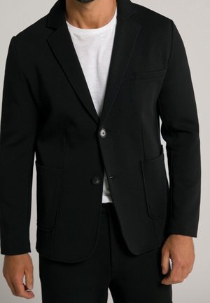 Uomo che indossa un blazer nero a due bottoni sopra una t-shirt bianca con scollo rotondo, con le mani rilassate ai lati su uno sfondo semplice.