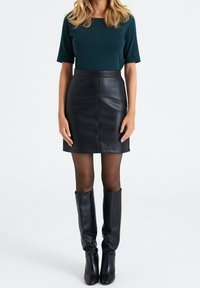 Black leather mini skirt with a high waist, paired with a dark green short-sleeve top. Model wears black knee-high boots.