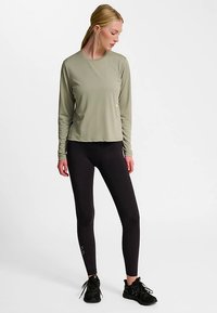 Langärmliges, olivgrünes Sportoberteil kombiniert mit schwarzen Leggings. Das Oberteil hat eine lockere Passform; die Leggings sind figurbetont mit einem Logodetail.