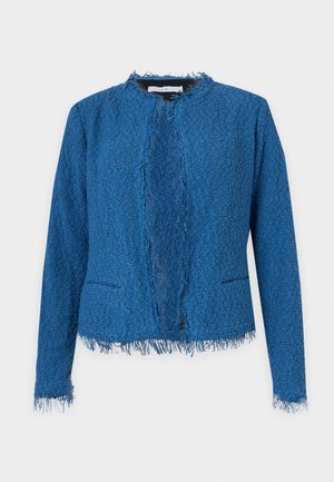 Frau steht mit einer blauen strukturierten Jacke, weißem Oberteil, weiten Jeans, Sandalen und hält eine große braune Tasche vor neutralem Hintergrund.