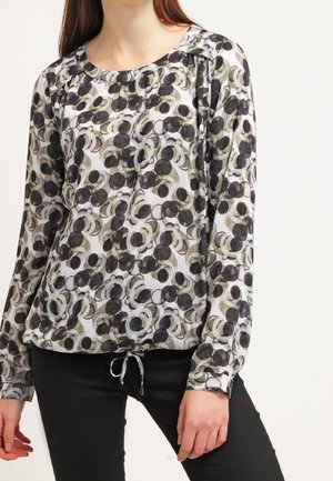 Kvinde iført bluse med lange ærmer med sort, gråt og beige cirkelmønster og sorte bukser, stående mod en ensfarvet hvid baggrund.